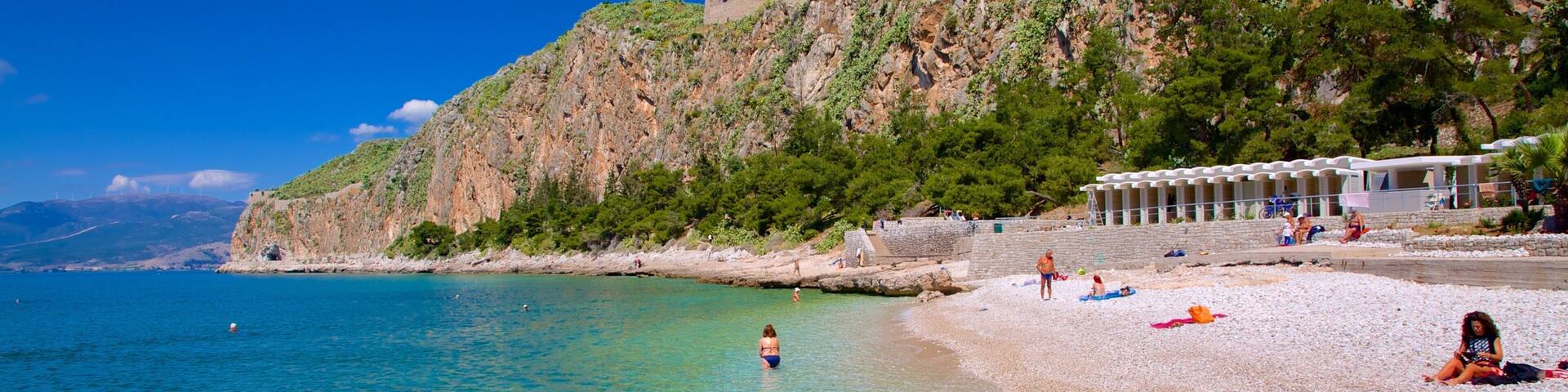 A pebble beach with clear turquoise water, a rocky cliff with greenery, and a building with a white roof.