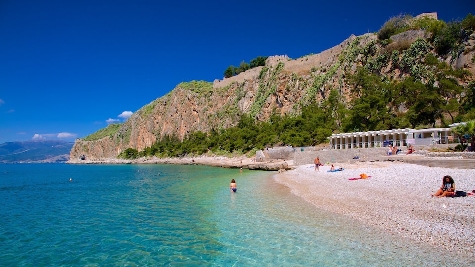 A pebble beach with clear turquoise water, a rocky cliff with greenery, and a building with a white roof.