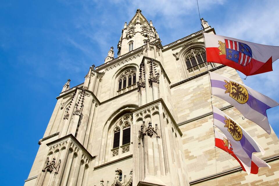 A historic cathedral with intricate Gothic architecture, flying flags, and a clear blue sky.