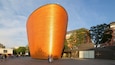 A large, orange, cylindrical building with a seagull perched on top.