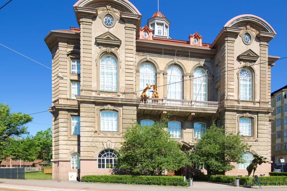 Um edifício histórico com grandes janelas em arco, uma varanda com esculturas de girafas e uma torre do relógio.