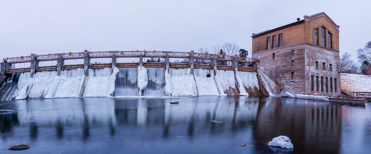 A dam with a wooden walkway, a brick building, and a frozen river.