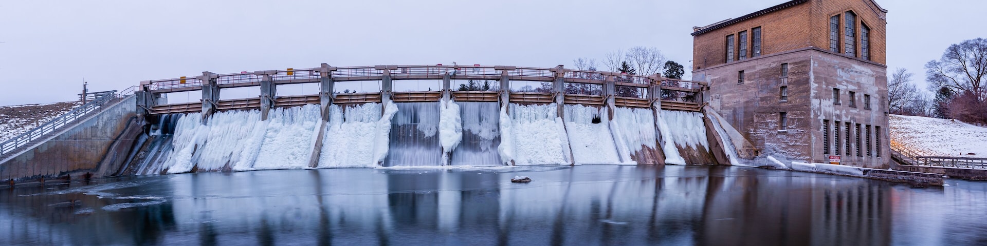 A dam with a wooden walkway, a brick building, and a frozen river.