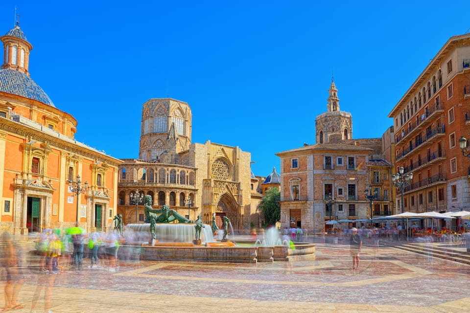A historic plaza with a fountain, ornate buildings, and a clear blue sky.