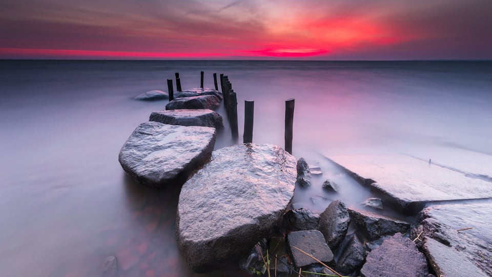 A rocky shoreline with a long pier extending into calm waters, under a vibrant sunset sky.