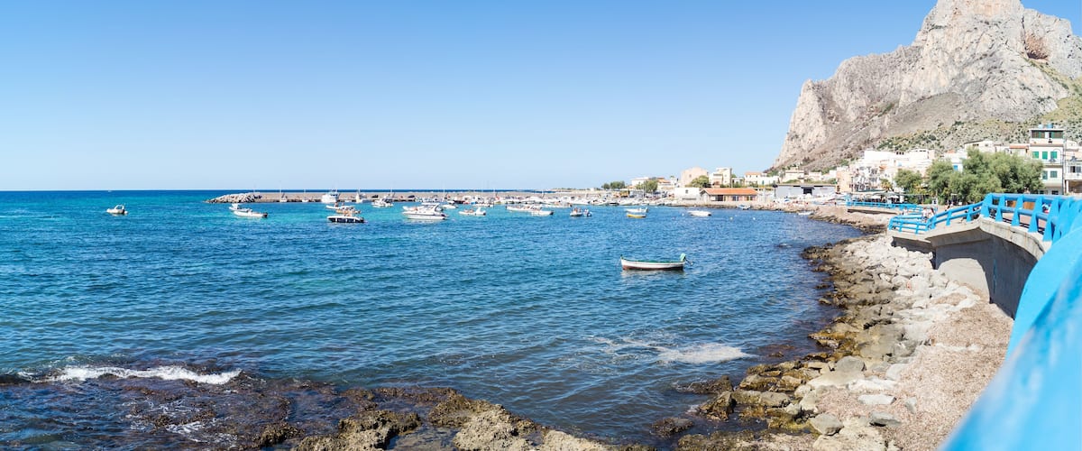 A coastal town with boats docked along the shore, a rocky outcrop, and buildings in the background.