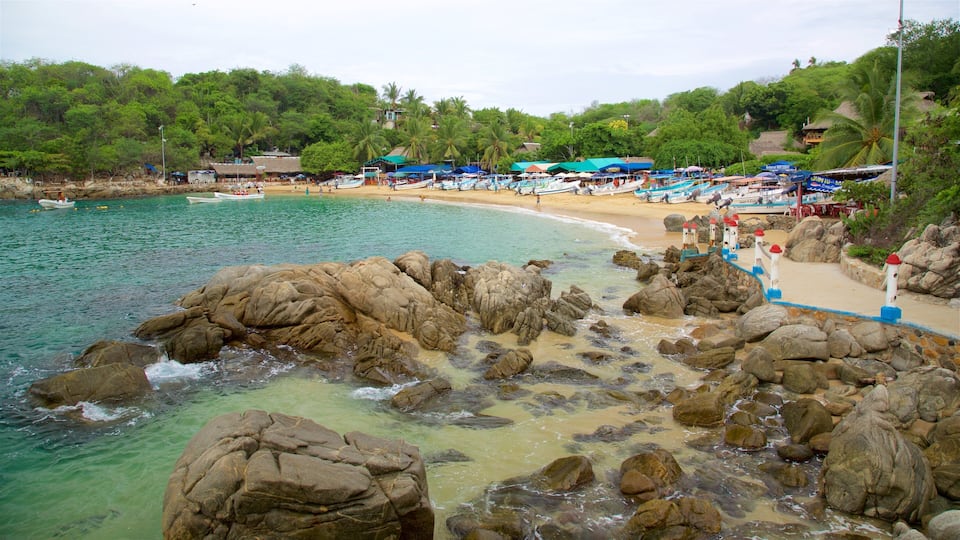 Una costa rocosa con agua turquesa cristalina, una playa arenosa y un telón de fondo de exuberantes árboles verdes y edificios con techos de paja.