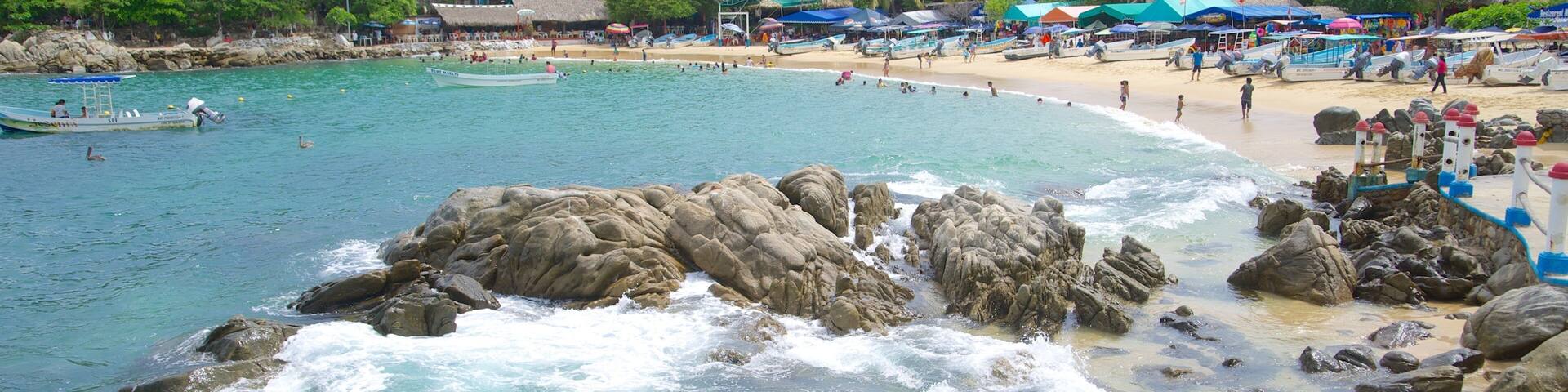 A beach with clear turquoise water, rocky shoreline, and lush green hills in the background.