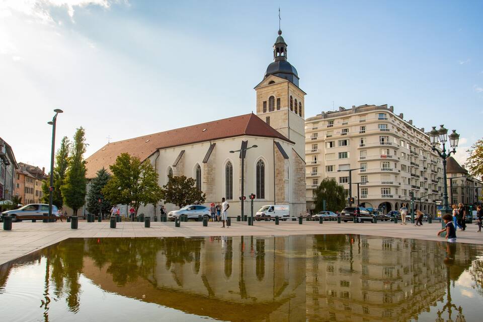 A church with a tower, a fountain reflecting the building, and surrounding urban buildings.