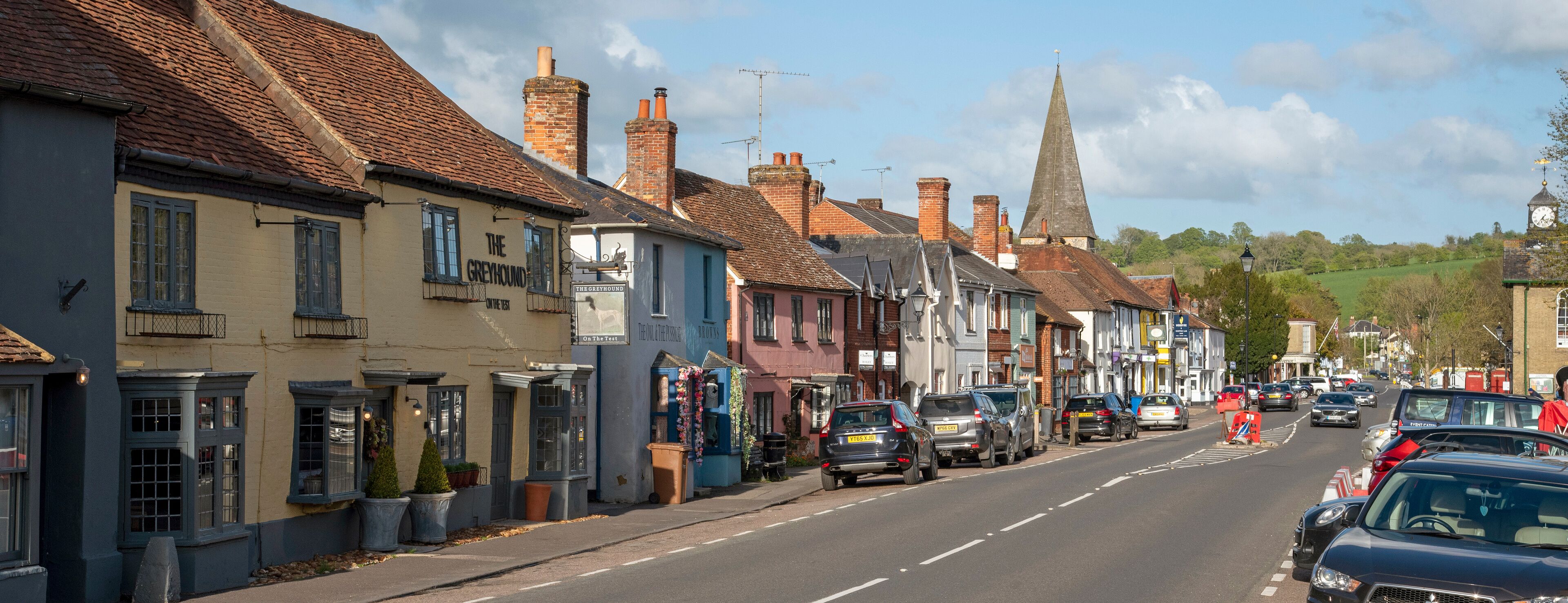 A street lined with historic buildings, a church spire, and parked cars.