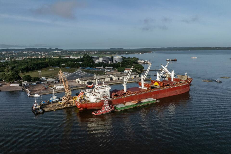 A large red cargo ship docked at a port with cranes and smaller boats in the background.