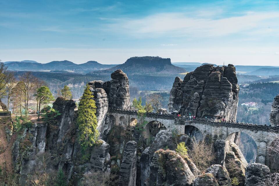 Eine Steinbrücke über einer schroffen Schlucht, im Hintergrund ein bewaldetes Gebirge.