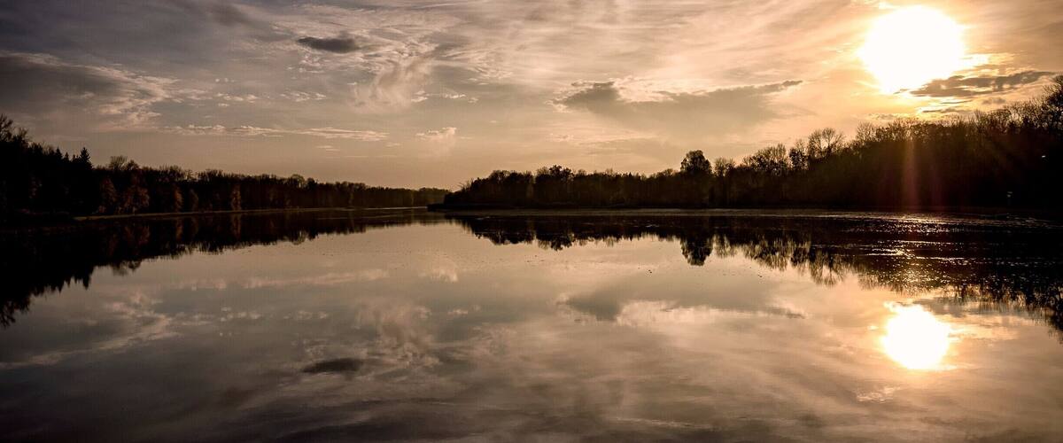 A calm lake reflecting the sky with the sun setting behind trees.
