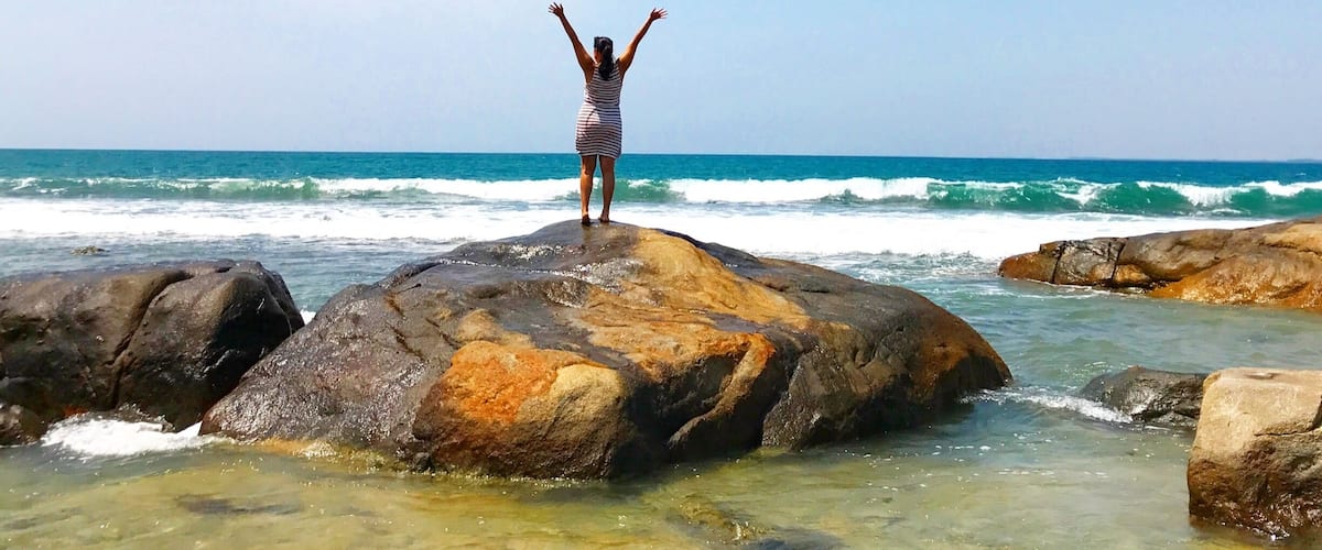 A person standing on a rock with arms raised, overlooking the ocean.