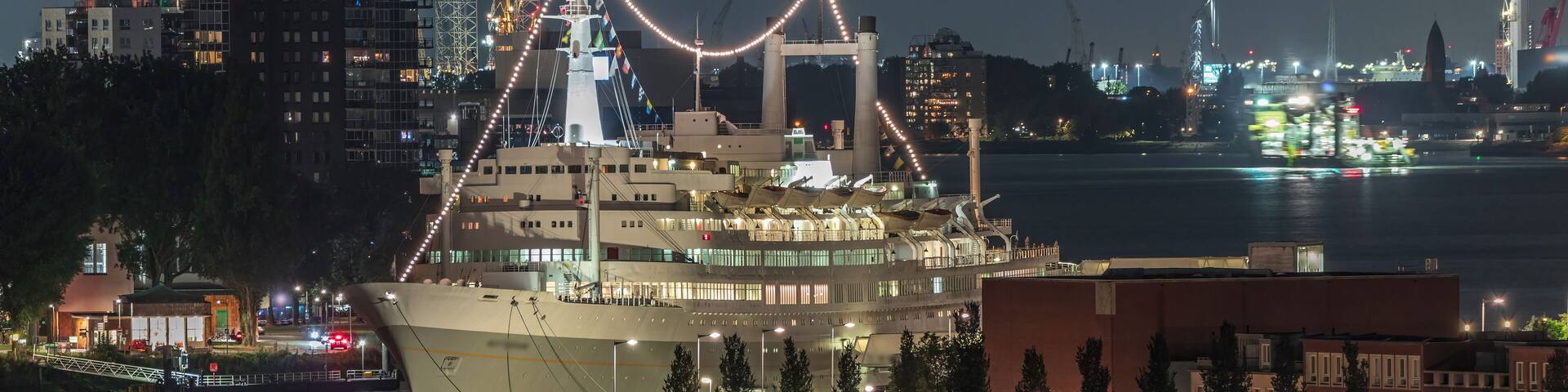 A large cruise ship docked at a port with city buildings and a river in the background.