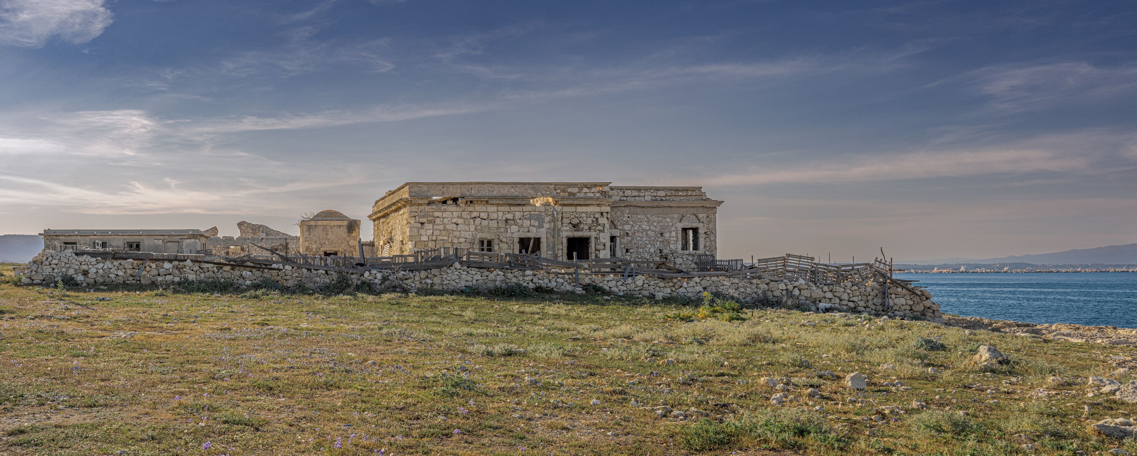An ancient stone building with a stone wall and a wooden fence, overlooking a body of water.