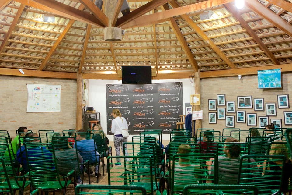Un espacio interior cubierto con sillas verdes, techo de madera, una pizarra con texto y una pantalla de proyector.