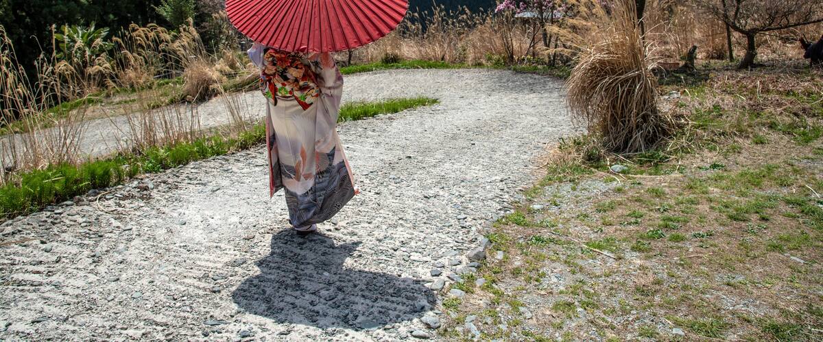 A person walking on a gravel path with a red umbrella, surrounded by trees and a mountainous landscape.