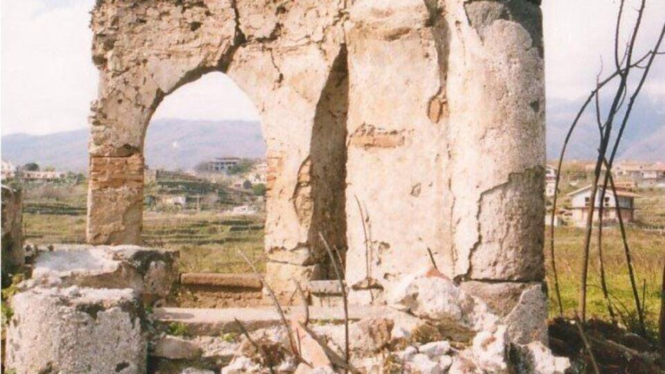 Ancient ruins with a large archway and columns, surrounded by rocks and vegetation.