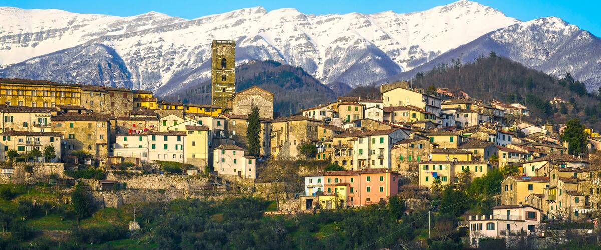 A historic town with colorful buildings, a prominent church tower, and snow-capped mountains in the background.