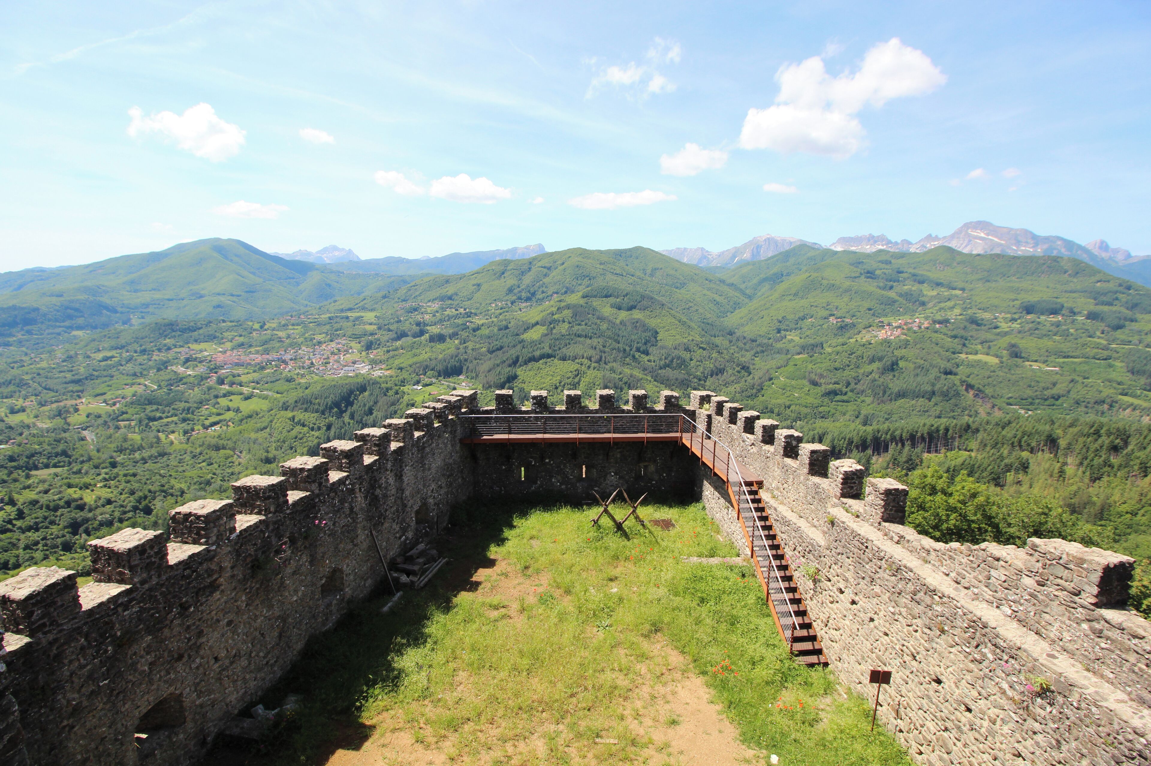 A historic stone fortress with a surrounding wall, set against a backdrop of lush green hills and distant mountains.