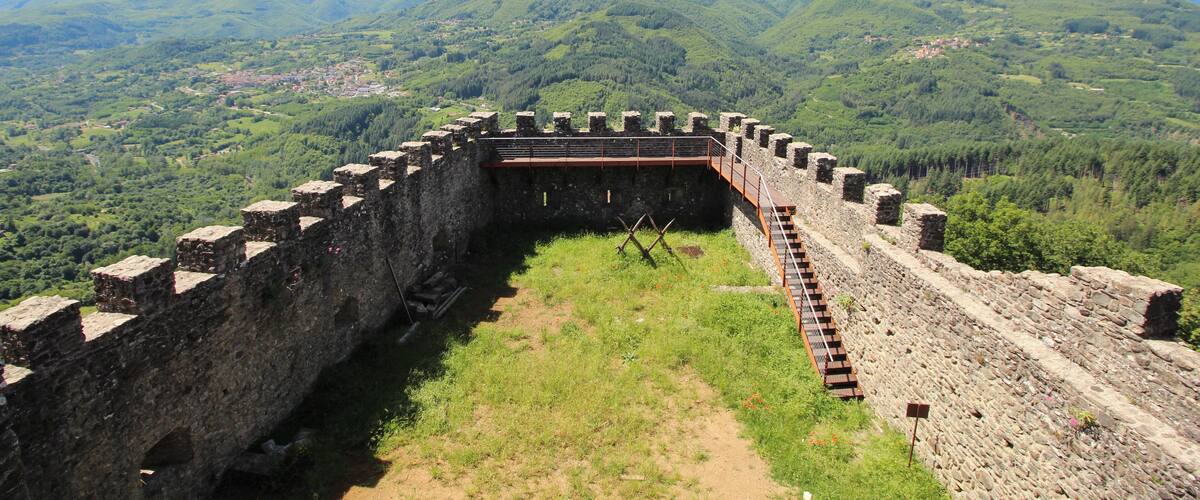 A historic stone fortress with a surrounding wall, set against a backdrop of lush green hills and distant mountains.