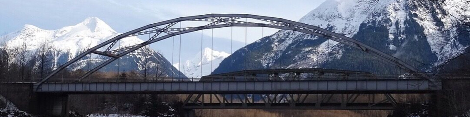 Eine Stahlbrücke über einem ruhigen Fluss, im Hintergrund schneebedeckte Berge.