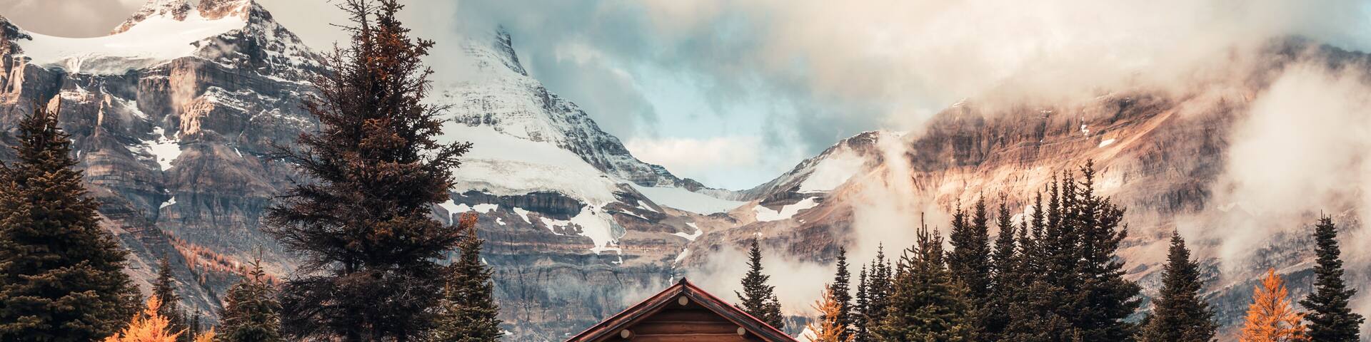 A log cabin surrounded by evergreen trees and a snow-capped mountain in the background.