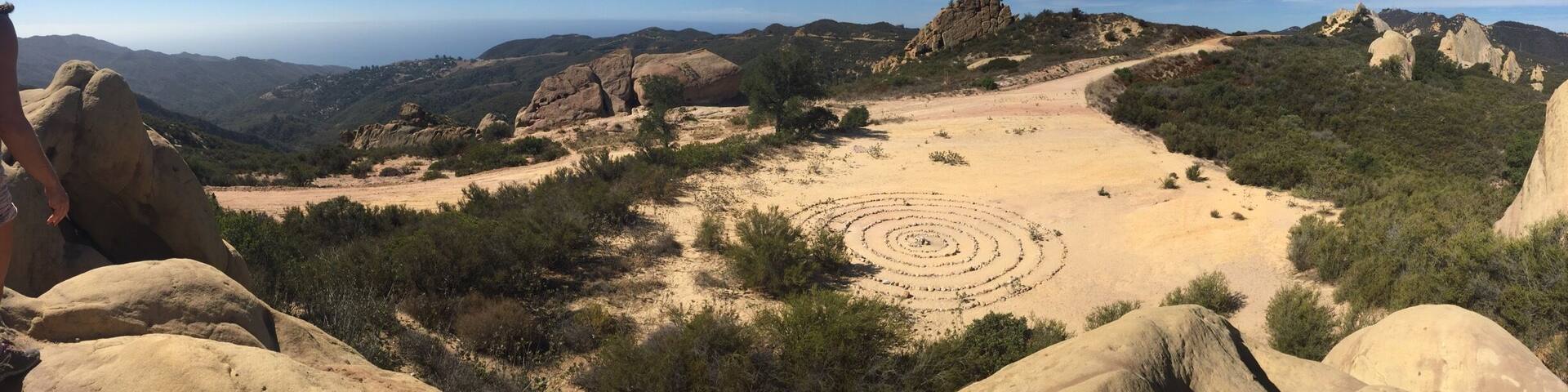 Paisaje desértico con una gran formación rocosa en primer plano, un patrón circular en el segundo plano y montañas distantes bajo un cielo despejado.