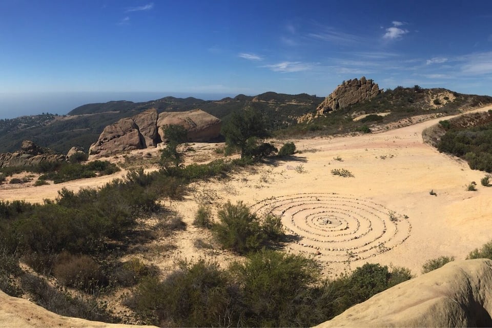 A desert landscape with a large rock formation in the foreground, a circular pattern in the middle ground, and distant mountains under a clear sky.