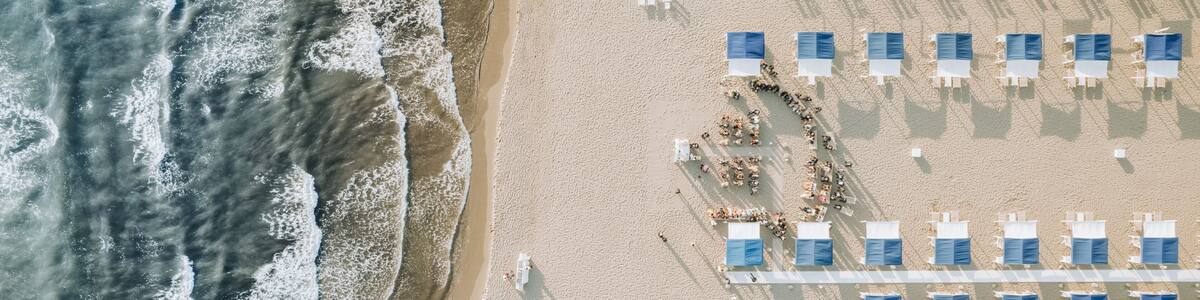 An aerial view of a beach with rows of sun loungers and a few people walking along the shore.