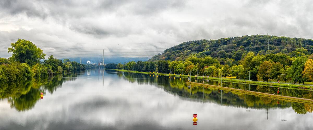 A calm river reflecting trees and a distant industrial facility.