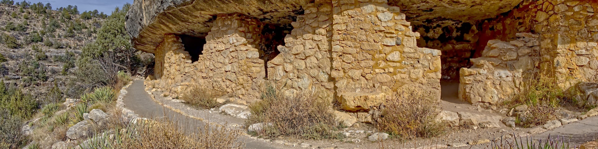 A stone ruin with a cave-like structure, surrounded by dry vegetation and a winding path.