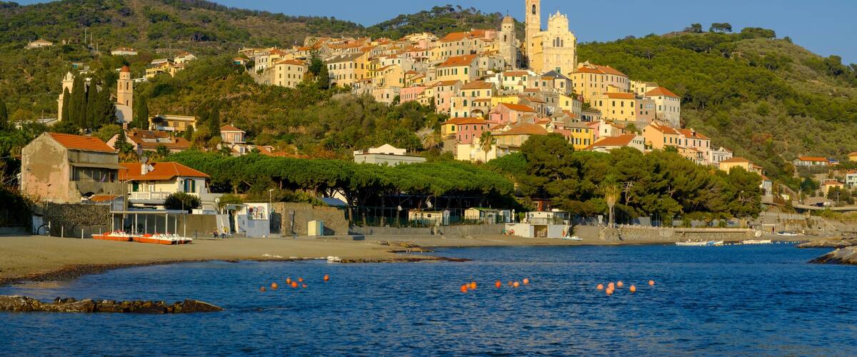 A coastal town with a prominent church on a hill, colorful buildings, and orange buoys in the water.