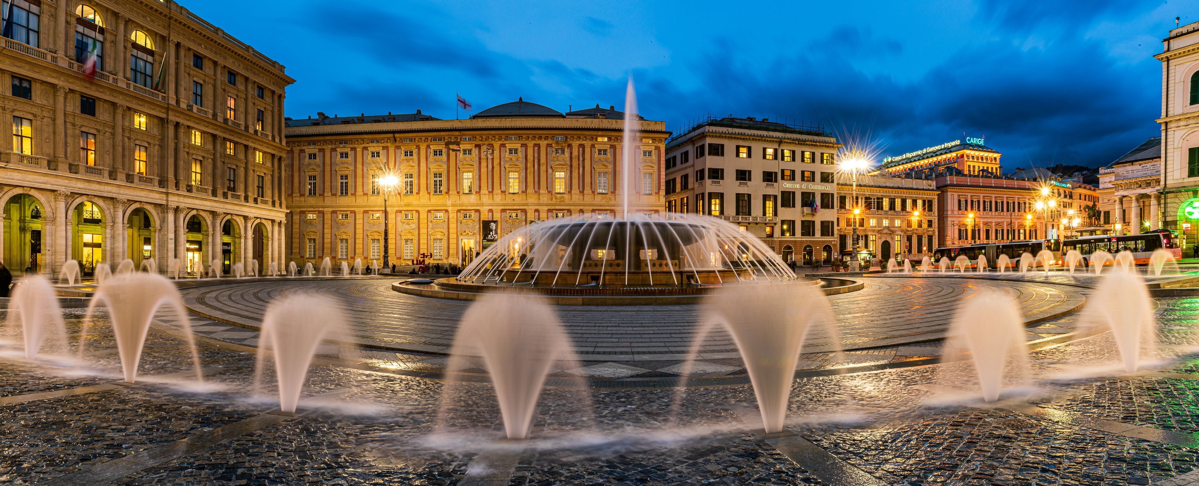 A historic square with a central fountain, ornate buildings, and a clear evening sky.