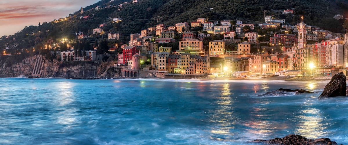 A coastal town with colorful buildings, a rocky shoreline, and a sunset sky.