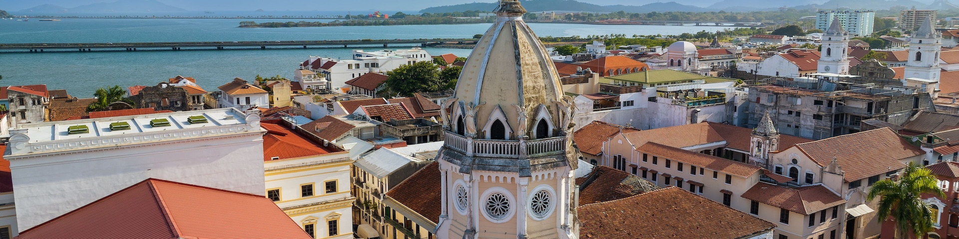 Paisagem costeira com um campanário de igreja imponente, um rio e montanhas ao longe.