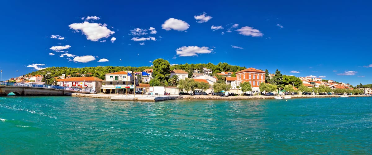 A coastal town with a bridge over turquoise water, buildings with red roofs, and a backdrop of green hills.