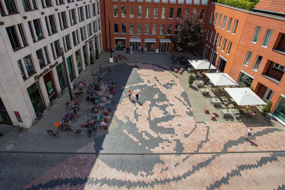 A plaza with a patterned pavement, surrounded by buildings with storefronts and outdoor seating areas.