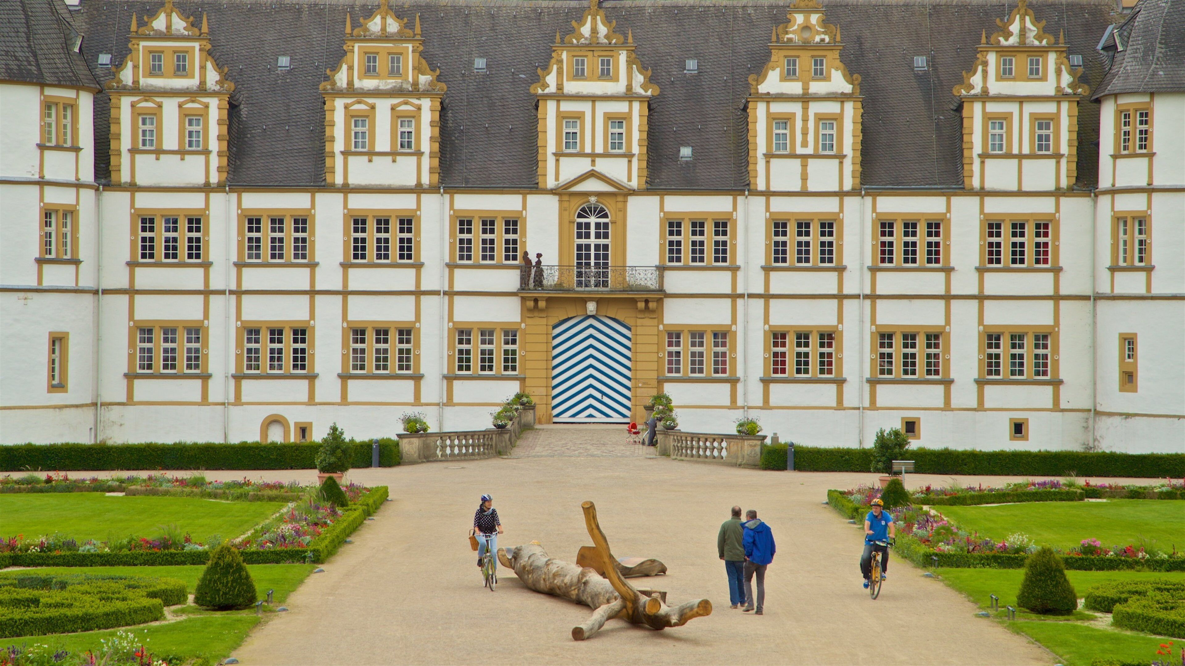 A large, ornate building with a symmetrical facade, a central entrance with a distinctive blue and white chevron pattern, and a well-manicured garden with hedges and flower beds.