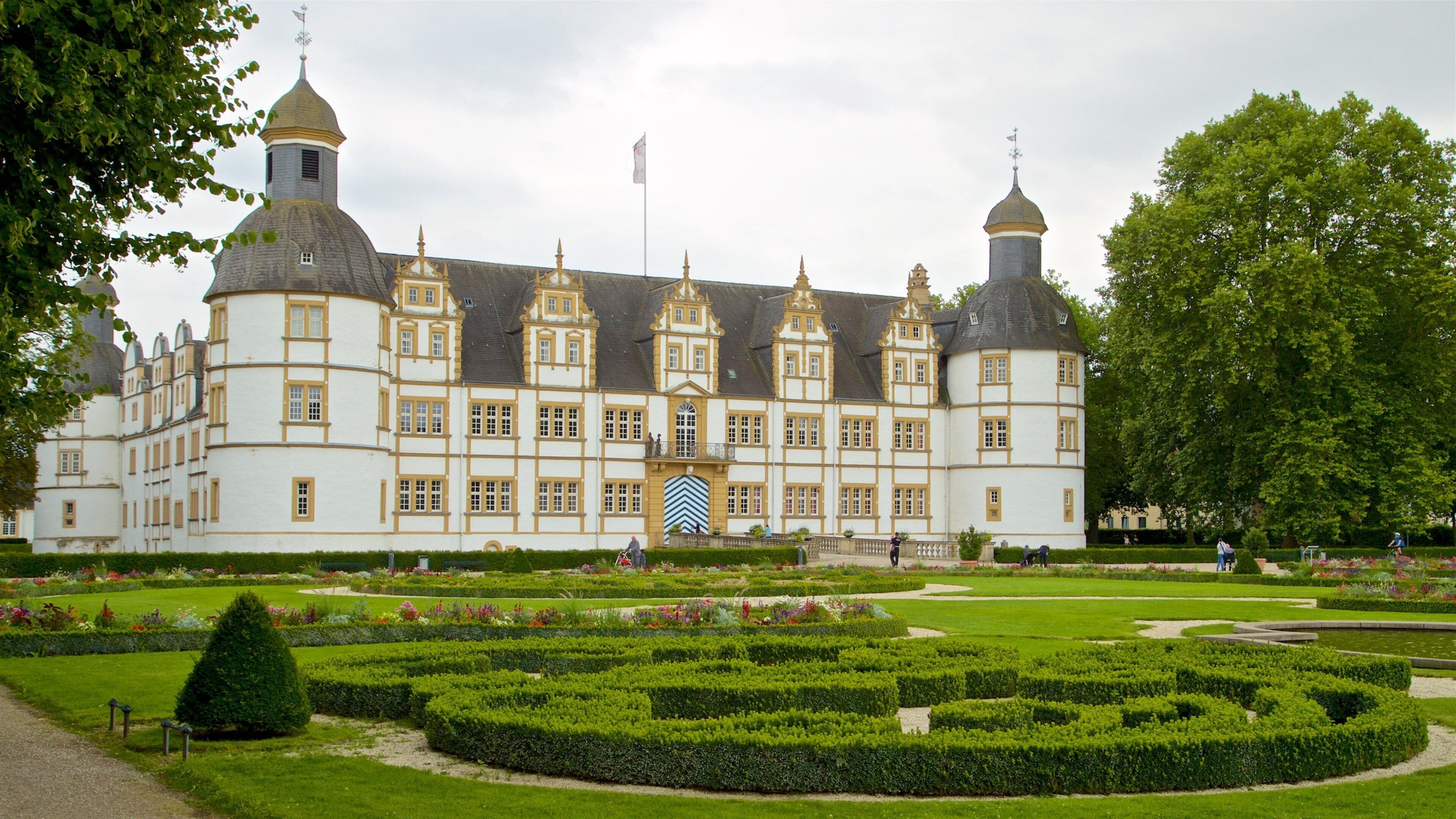 A large, historic building with multiple towers and a well-maintained garden with hedges and flowers.