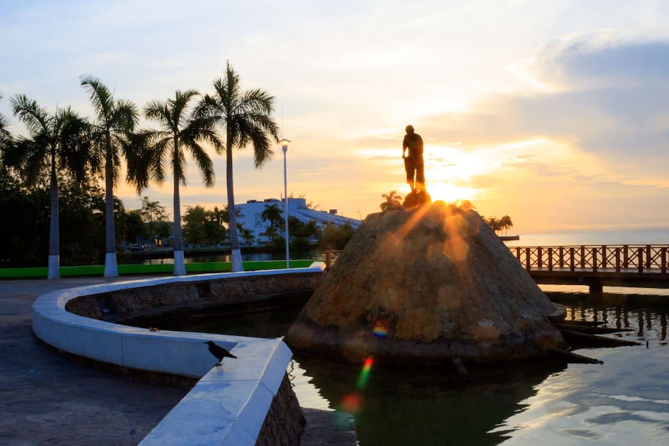 A sunset over a water feature with a statue on a rock, palm trees, and a bridge in the background.