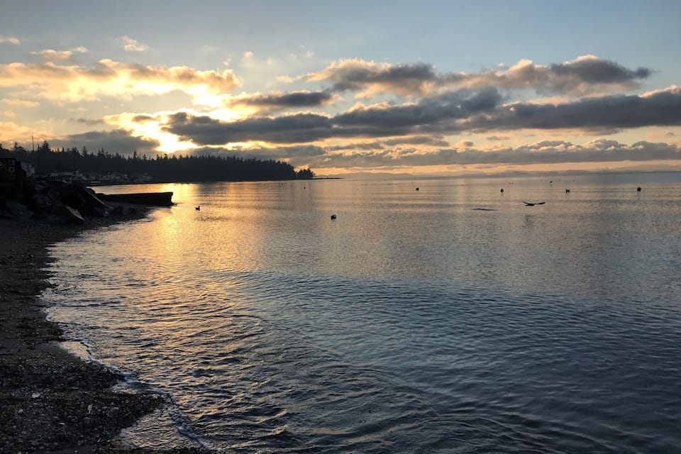 A calm waterfront at sunset with a clear sky, a distant shoreline, and a few buoys in the water.