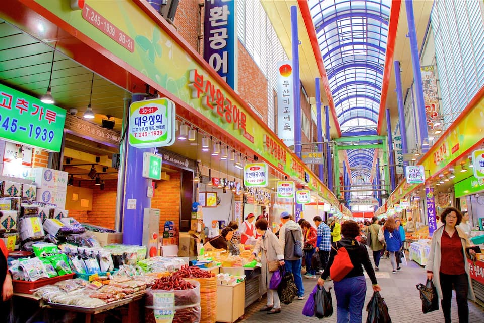 Un mercado interior concurrido con varios puestos que ofrecen comida y productos, y letreros que muestran precios y ubicaciones.