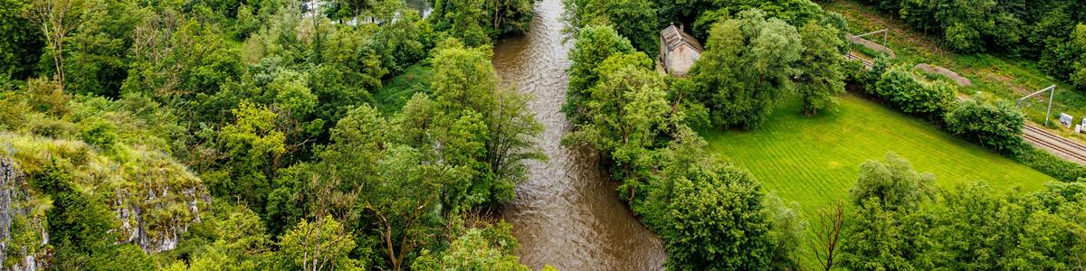 Valley with abundant green leafy trees and Ourthe river with abundant water slightly overflowing, mountains with horizon in background, seen from Logne castle, cloudy day in Ferrieres, Belgium
