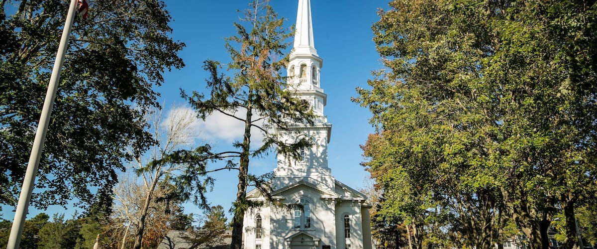 A white church with a tall steeple, surrounded by trees and a cemetery.
