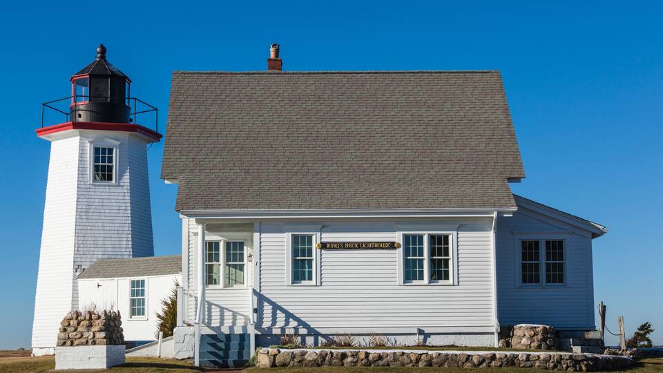 A white lighthouse with a black top and a white building with a sign reading "Wings Neck Lighthouse".