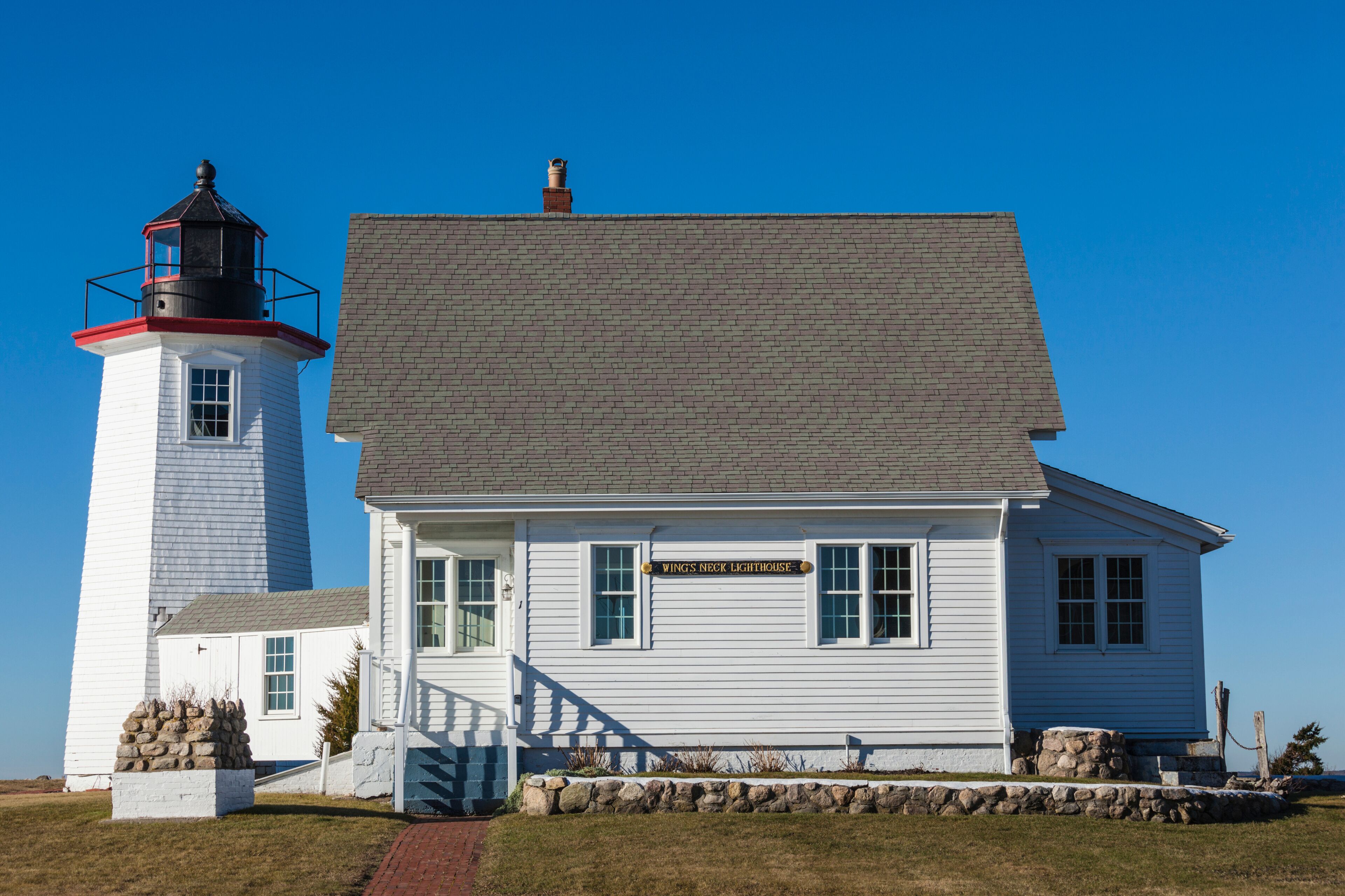 A white lighthouse with a black top and a white building with a sign reading "Wings Neck Lighthouse".