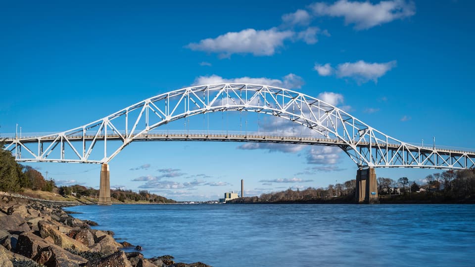 A large steel bridge spanning a river with rocky shores and trees on one side.