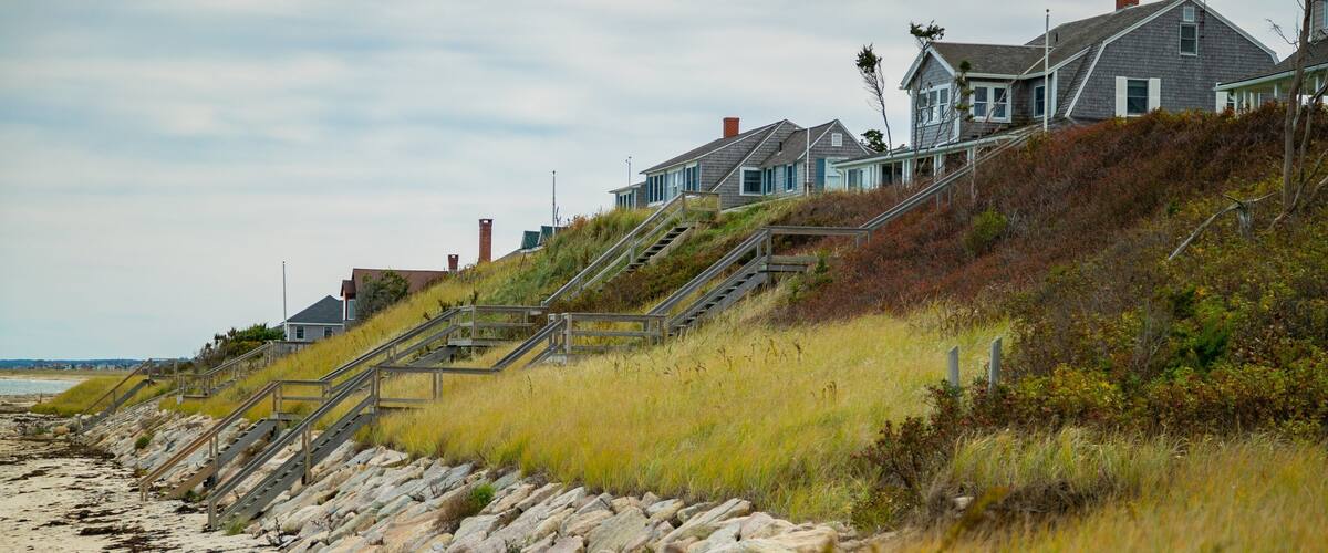 A coastal landscape with a beach, grassy dunes, and a row of houses with stairs leading up to them.